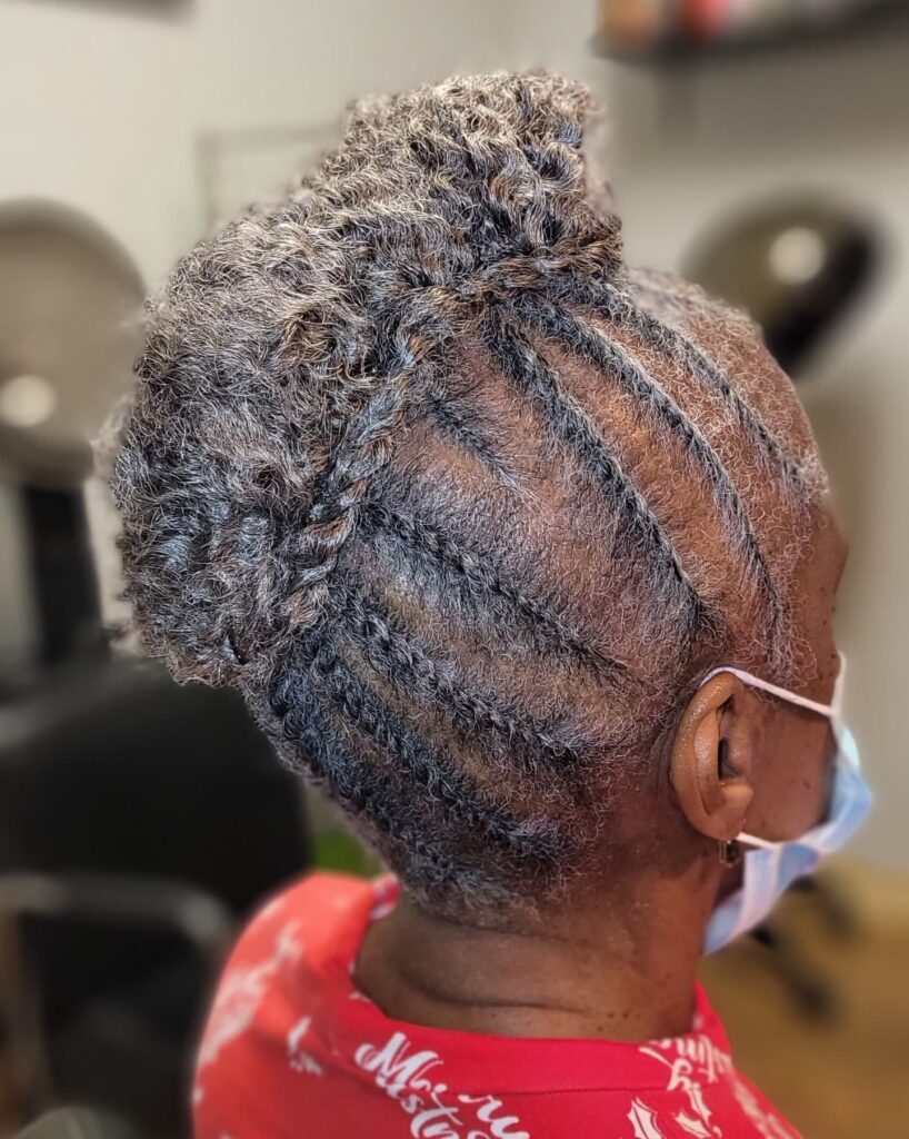 An older woman viewed from the side, featuring an updo with side cornrows and a voluminous curly bun in silver and charcoal grey.