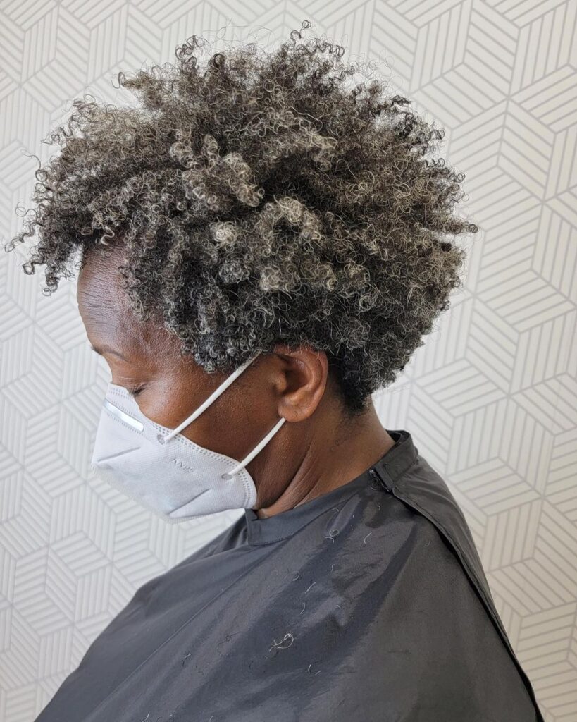 A woman in a salon cape showcasing a voluminous short afro with salt-and-pepper curls styled in a layered shag.