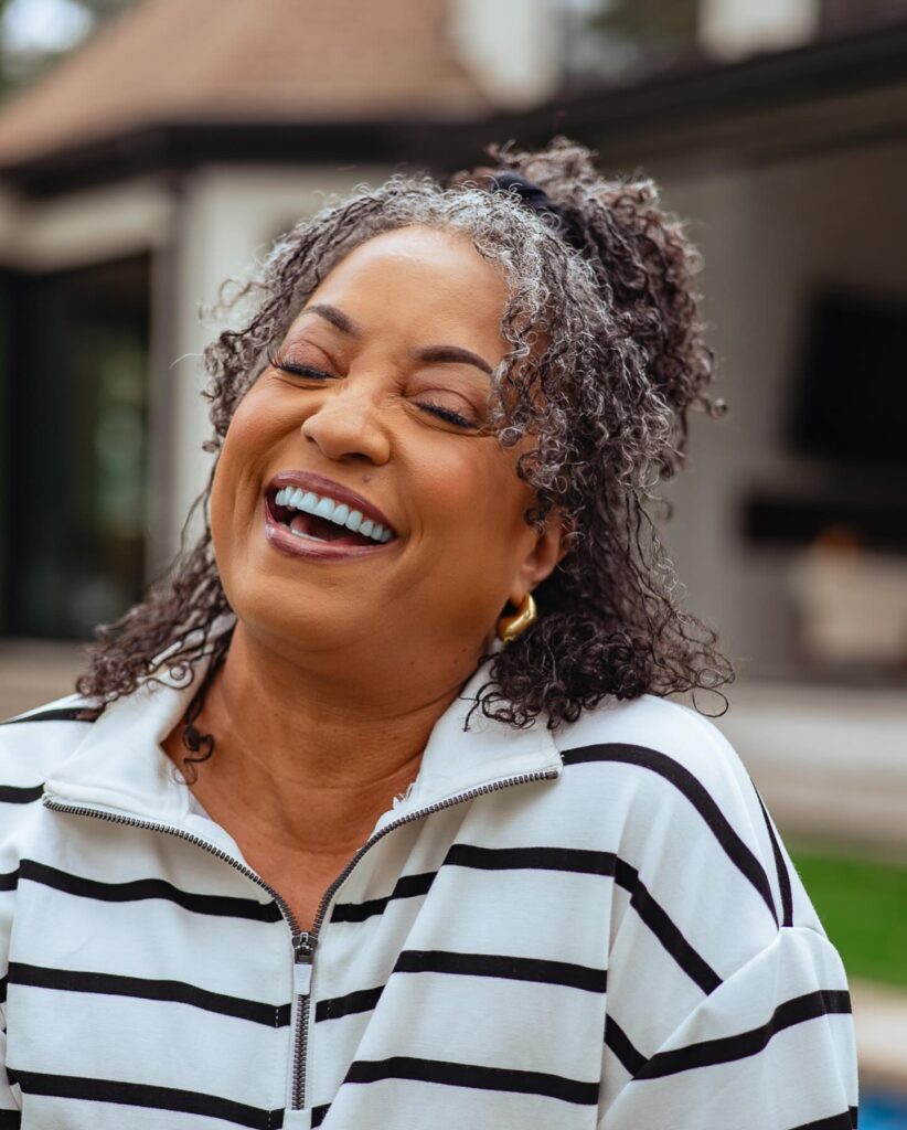 A laughing woman wearing her curly salt-and-pepper hair in a half-up style with face-framing ringlets.