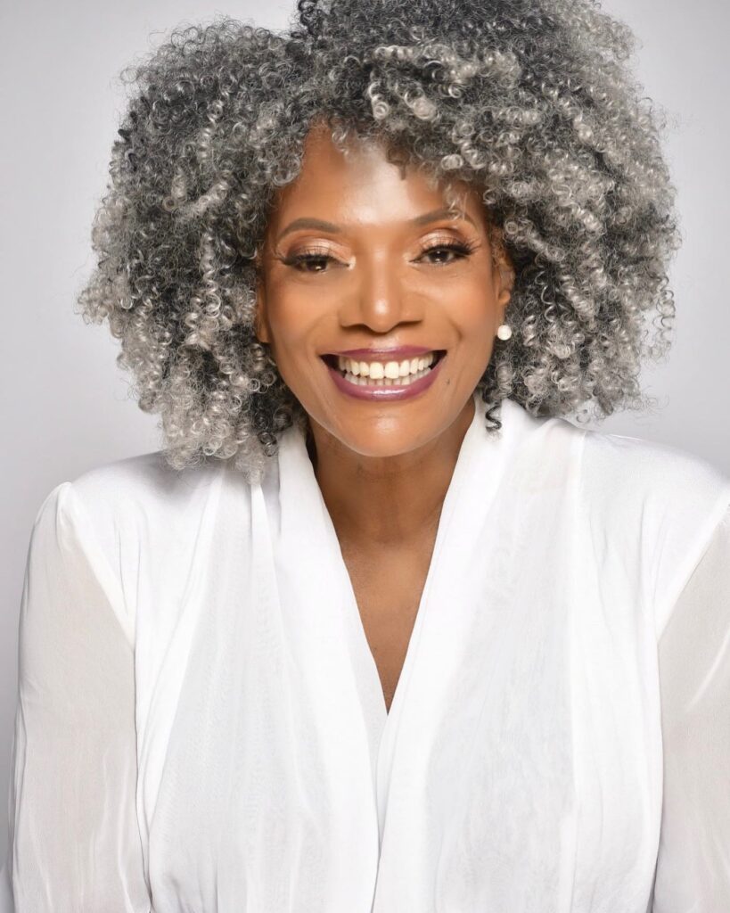 A smiling woman with a voluminous, medium-length rounded afro in a vibrant salt-and-pepper grey shade.