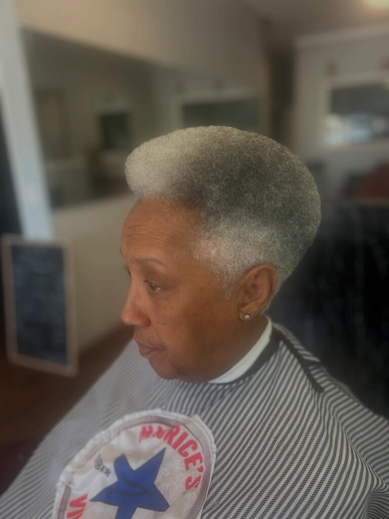 A woman in a barber chair is showcasing a precision tapered fade with short grey hair and a rounded top.