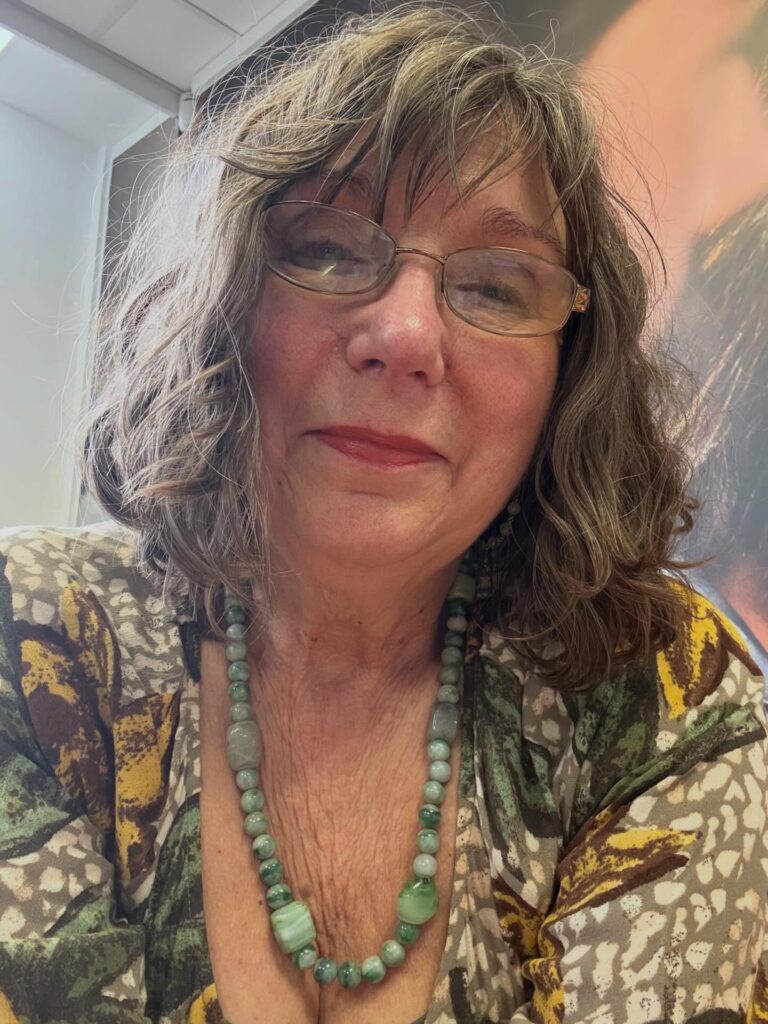 A woman over 60 with glasses wearing a textured shoulder-length wavy hairstyle with wispy bangs and blonde highlights.