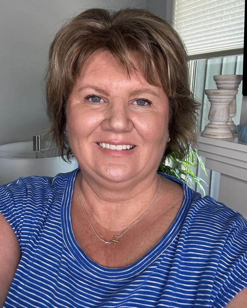 A smiling woman over 60 with a voluminous ash brown feathered shag haircut featuring piecey bangs and flipped-out ends.