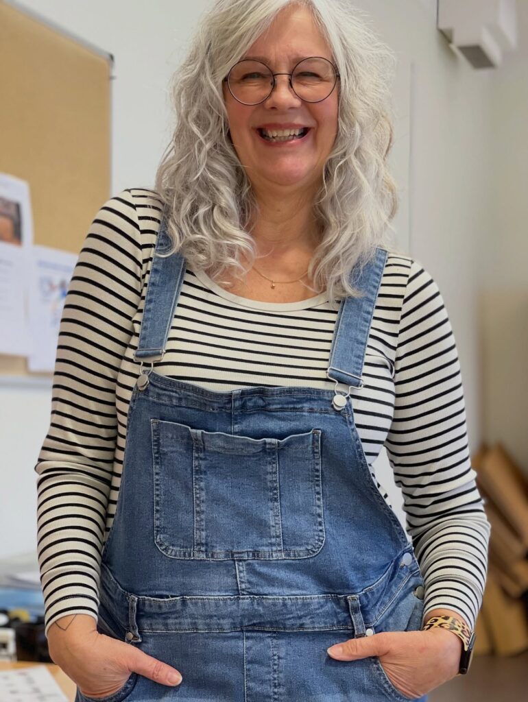 A smiling woman over 60 with long wavy silver hair wearing denim overalls and a striped shirt.