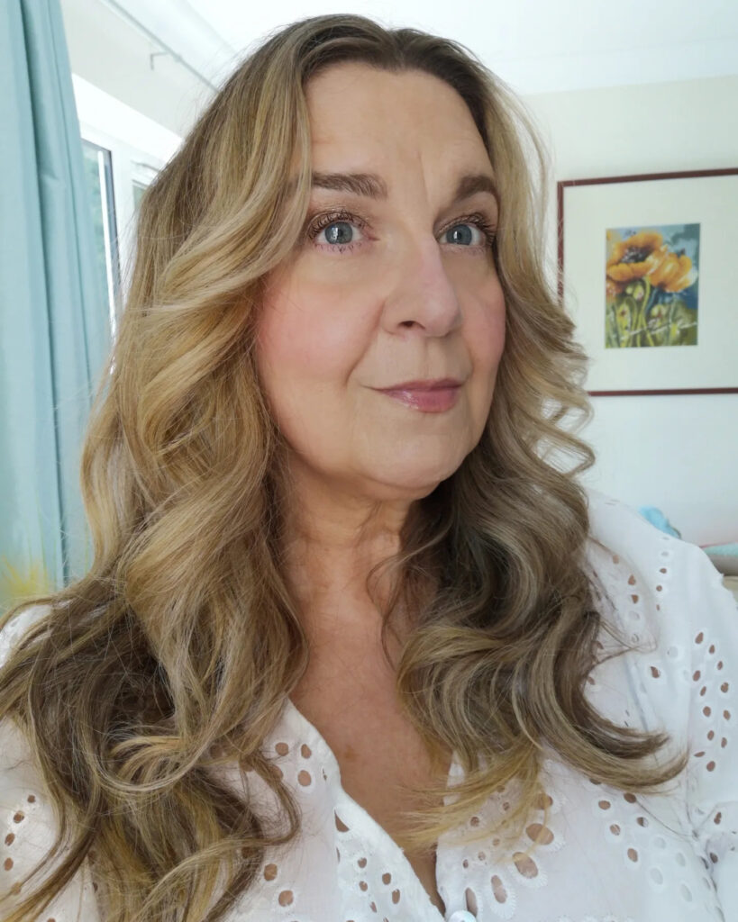 A woman over 60 with long blonde balayage hair styled in voluminous cascading waves.