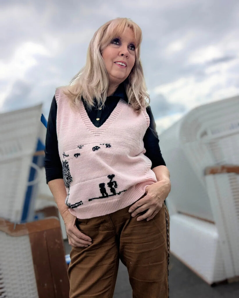 A woman over 60 with long layered champagne blonde hair and bangs, wearing a pink sweater vest and brown trousers.
