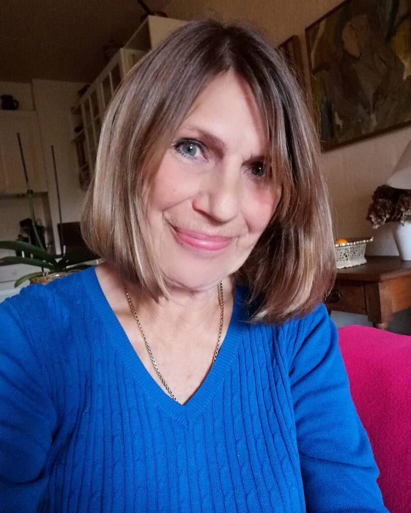 A woman with a sleek honey-blonde bob hairstyle smiling while wearing a blue V-neck sweater.