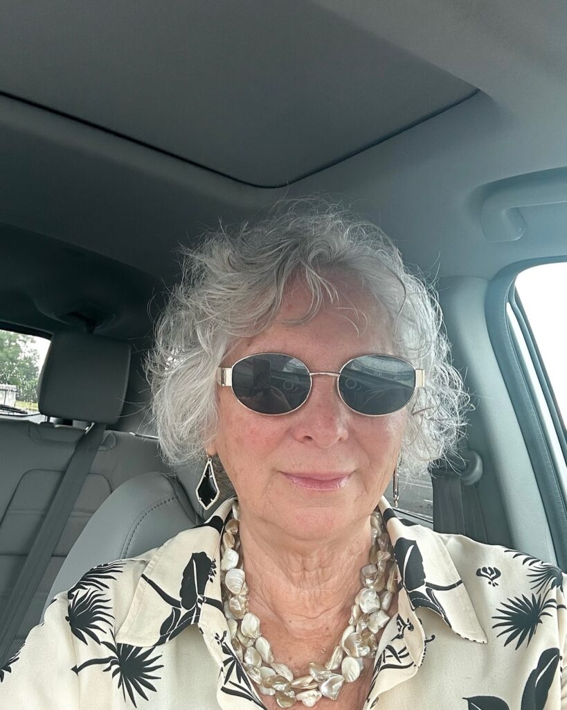 A woman with short curly silver hair wearing sunglasses and a patterned shirt is sitting in a vehicle.