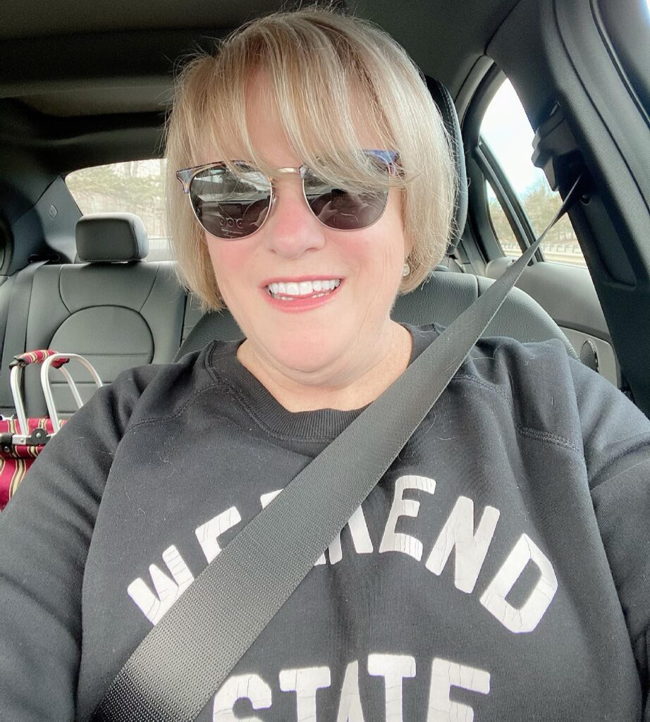 A woman over 60 with blonde hair in a rounded jaw-length bob and wispy bangs smiling in a car.