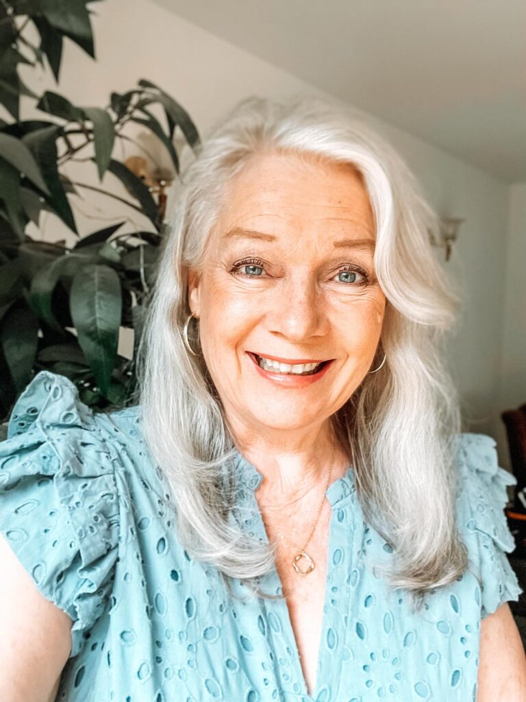 A smiling woman over 60 with long silver hair styled with a deep side part and soft, inward-curving layers.