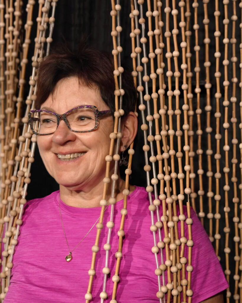 A smiling woman over 60 with a short spiky auburn pixie cut wearing purple glasses and a pink shirt.