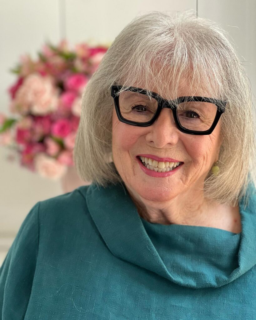 A smiling woman over 60 with black glasses wearing a classic silver chin-length bob with full straight bangs.
