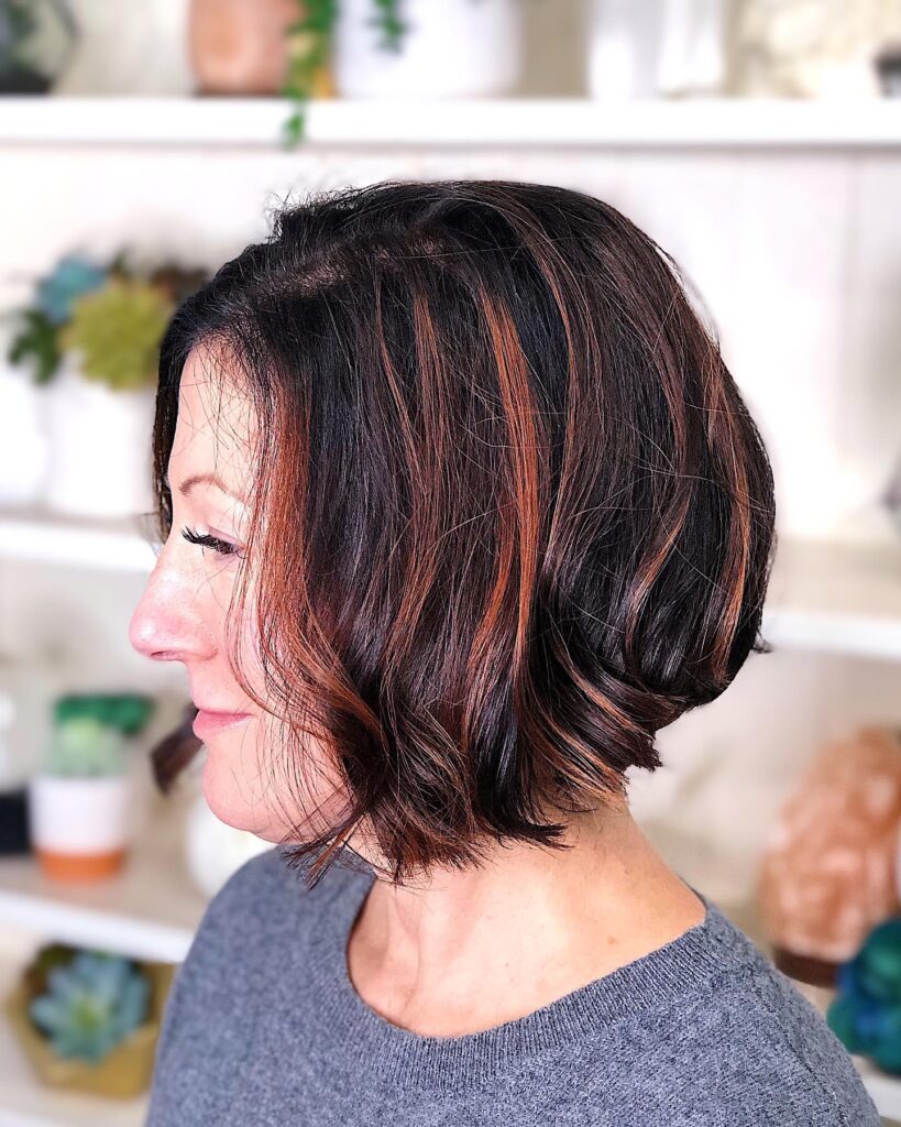 A woman over 60 shown from a side profile wearing a stacked A-line bob with dark brown hair and bright copper highlights.