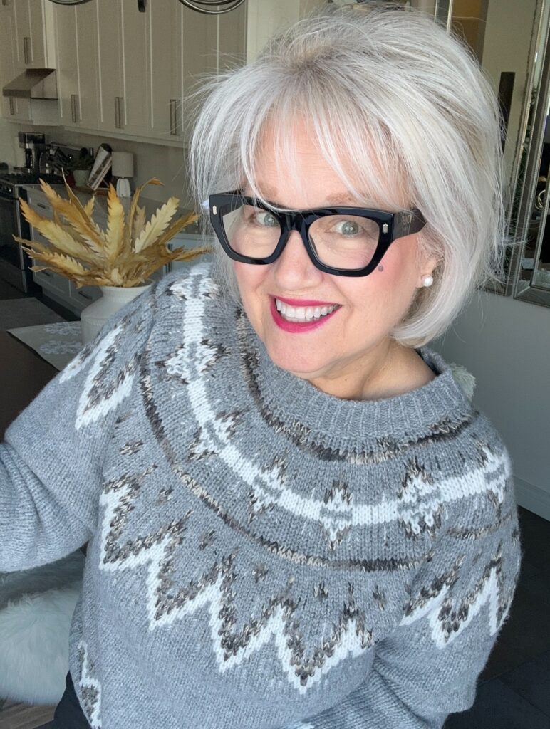 A smiling woman with a layered pearl-white bob and wispy bangs, wearing bold black glasses and a grey patterned sweater.
