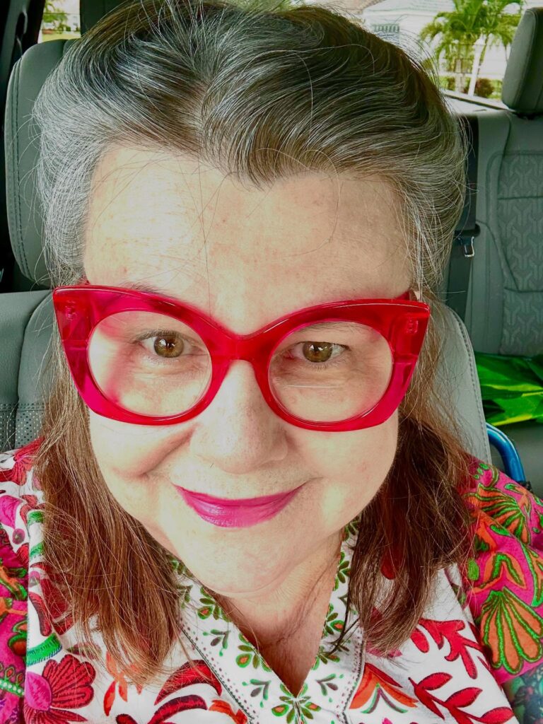 A woman with brown hair and silver roots styled in a vintage sweep, wearing bright red glasses.