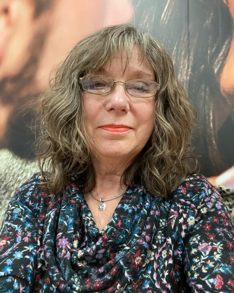A woman with glasses wearing a shoulder-length ashy brown shag haircut with soft curls and thin wispy bangs.