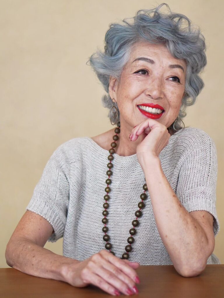A smiling woman with a short curly haircut styled in a unique dusty blue color.