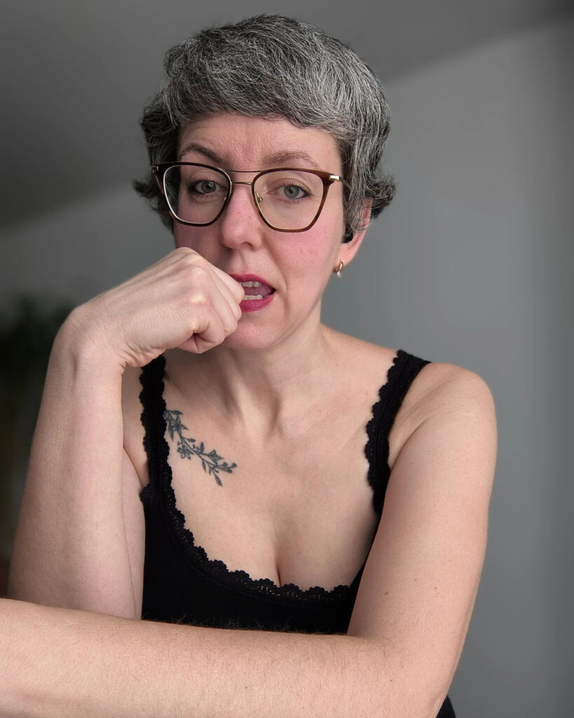 A woman with glasses and a tattoo wearing a short textured charcoal gray pixie cut with voluminous side-swept layers.