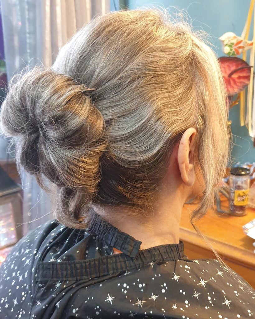 A rear view of a woman with silver hair styled into a low textured chignon bun with soft, loose curls framing her neck.