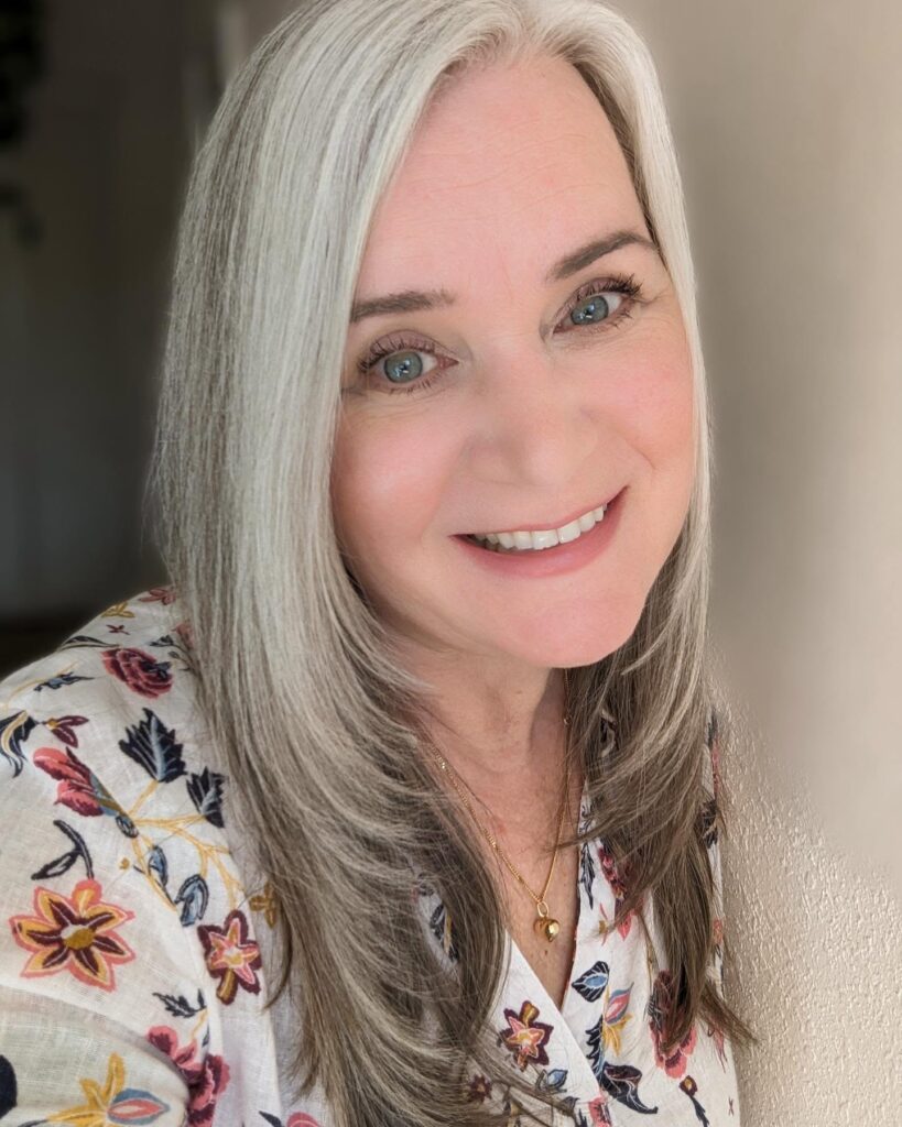 A smiling woman with long, straight silver hair featuring soft face-framing layers and a side part.