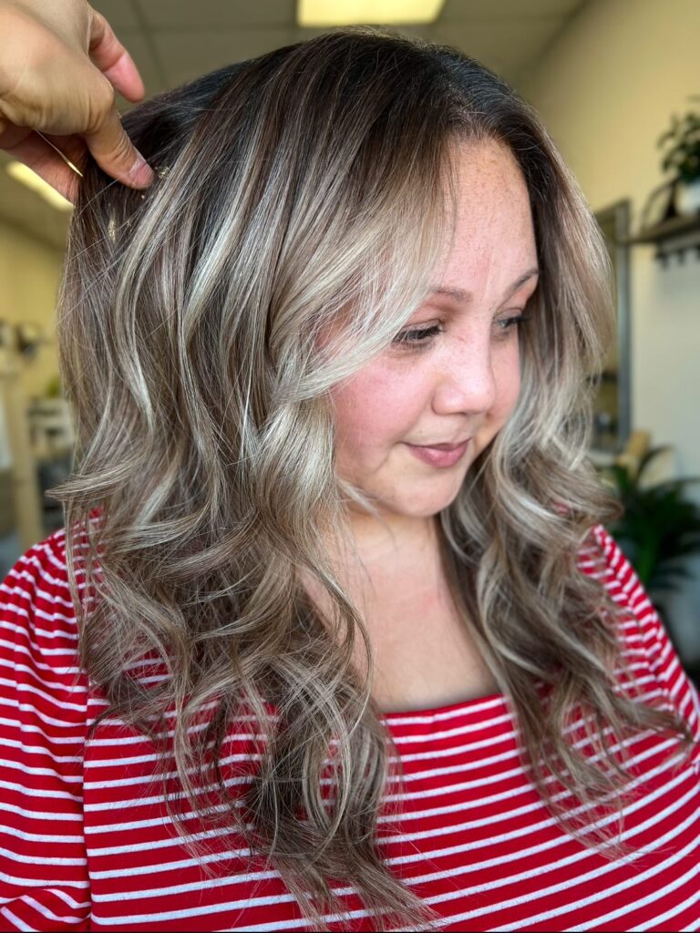 A woman with long, wavy brunette hair featuring cool ashy blonde balayage highlights and face-framing layers.