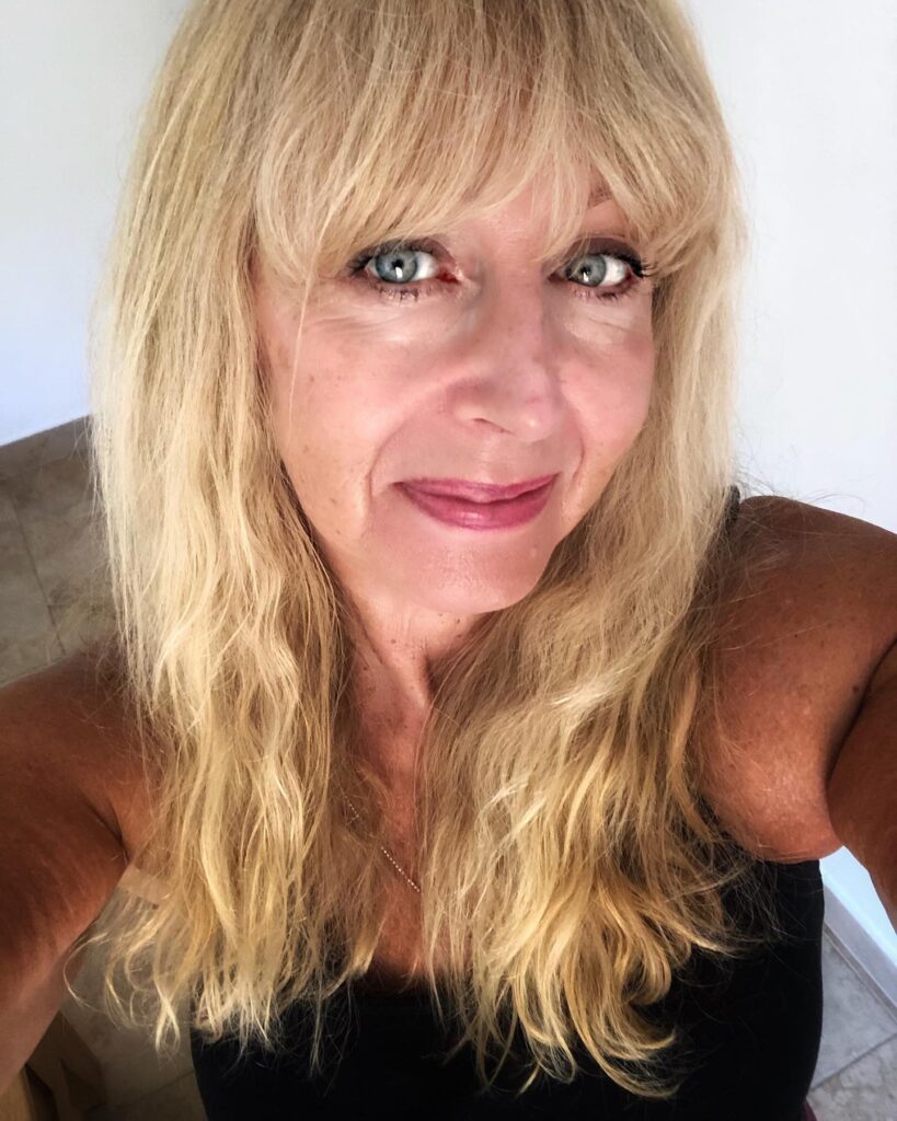 A woman with long, wavy, golden-blonde hair and a full, textured fringe.