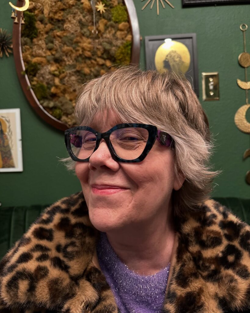 A woman with a short ash-blonde shaggy haircut featuring flicked-out layers and dark-framed glasses.
