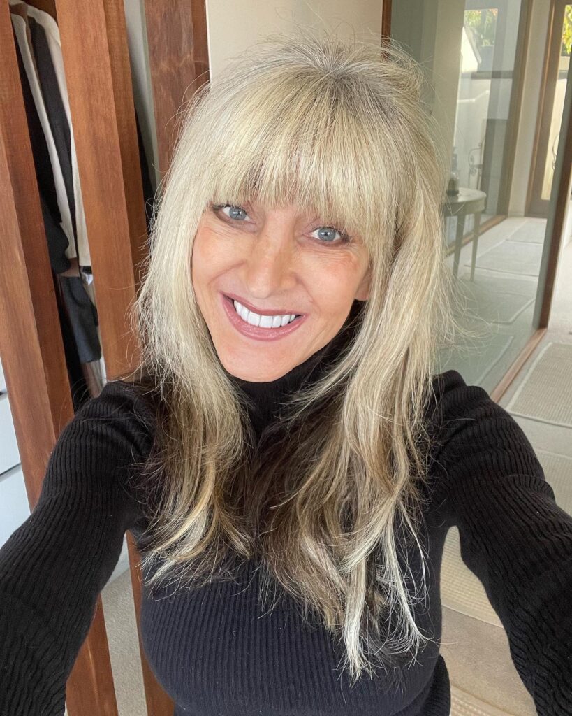 A woman over sixty with long ash-blonde hair featuring full straight bangs and feathered layers.