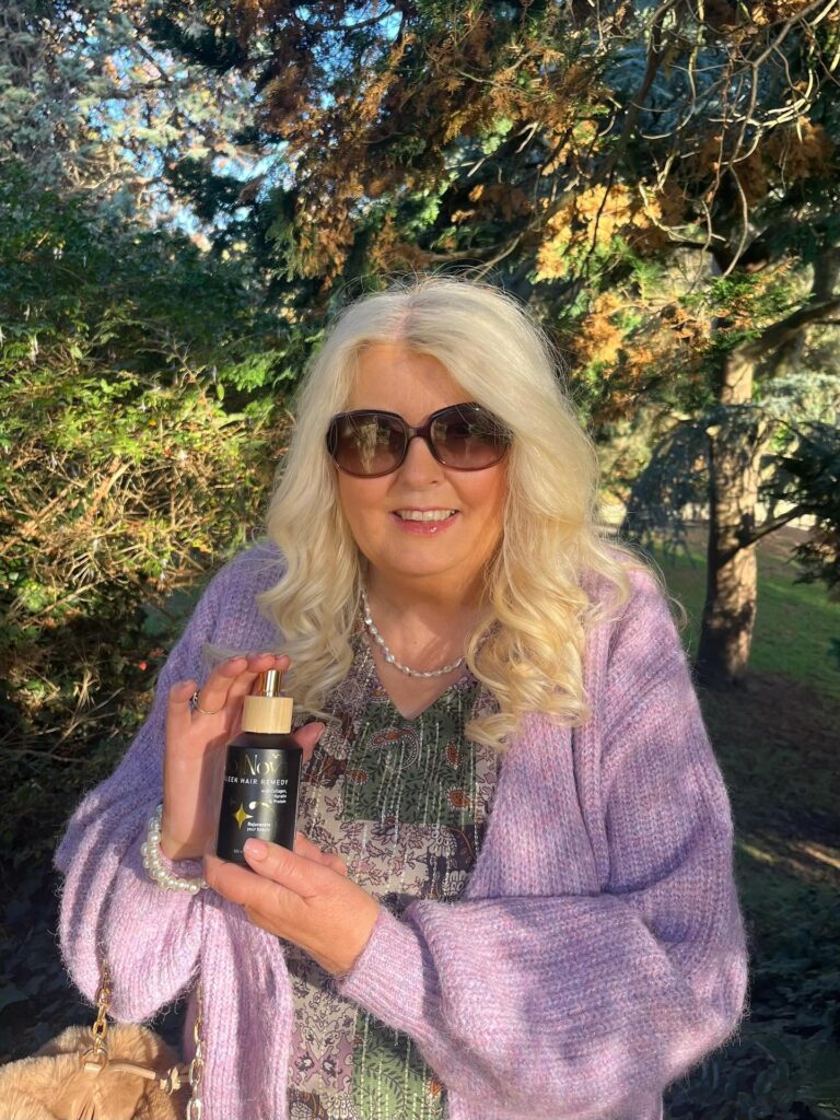 A woman with long, wavy butter-blonde hair wearing sunglasses and a purple cardigan.