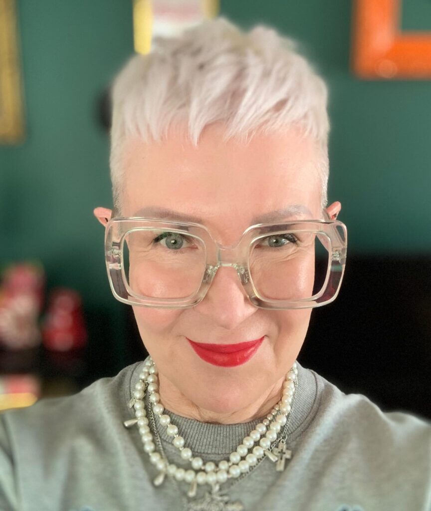 A woman over sixty wearing clear glasses and rocking an ultra-short platinum blonde pixie cut with textured styling on top.