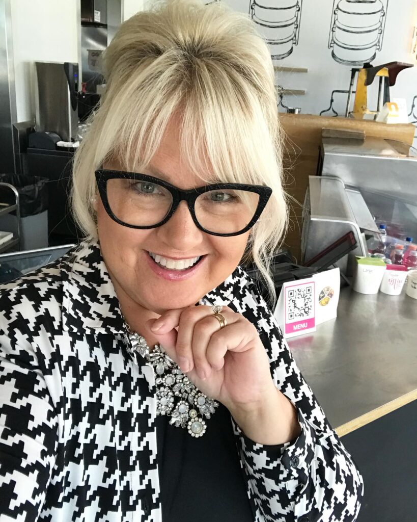 A woman with a vanilla blonde high updo and wispy bangs, wearing thick black glasses and a houndstooth jacket.