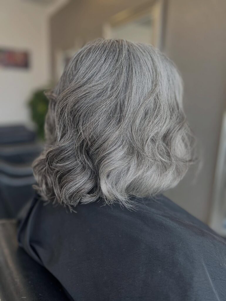 A rear-view photograph of a woman with a neck-length bob styled in soft waves, showcasing multi-dimensional silver and gray hair color.