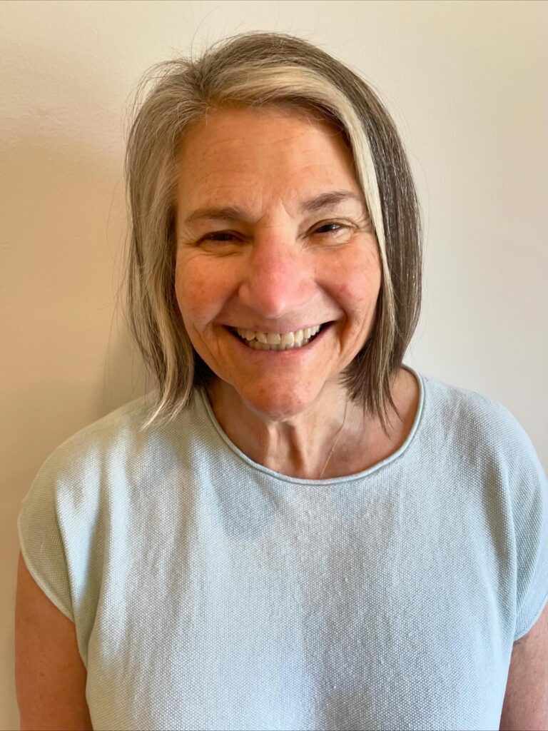 A smiling woman with a chin-length straight bob featuring natural salt and pepper hair and a prominent silver highlight at the front.
