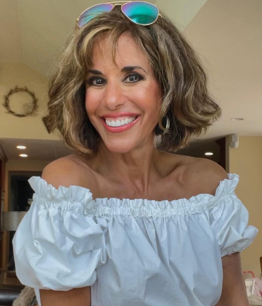 A smiling woman wearing a textured chin-length bronde bob with soft waves and wispy bangs.