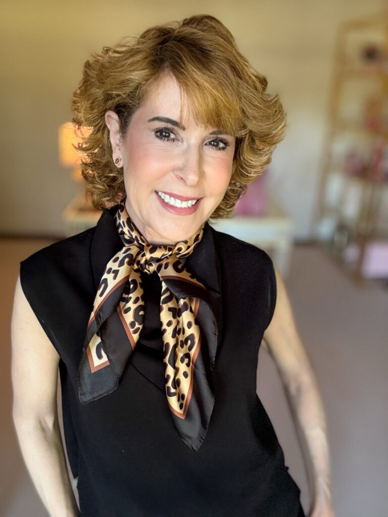 A smiling woman with a chin-length curly honey blonde bob featuring straight bangs and a lot of volume.