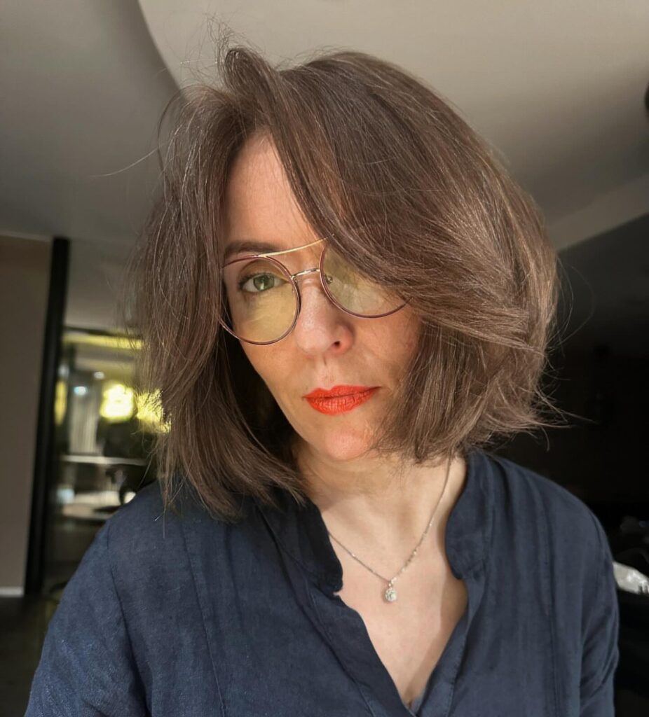 A woman wearing glasses with a voluminous shoulder-length lob in a deep mocha brown color, featuring a deep side part.