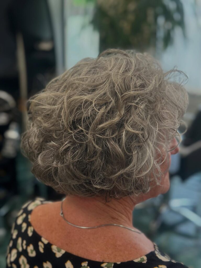 A rear side view of a woman with a voluminous chin-length bob featuring natural salt and pepper curls and layered texture.