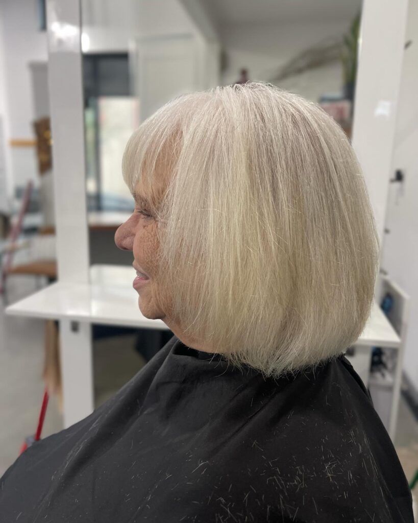 A side profile of a woman with a sleek chin-length platinum blonde bob and soft, wispy bangs.