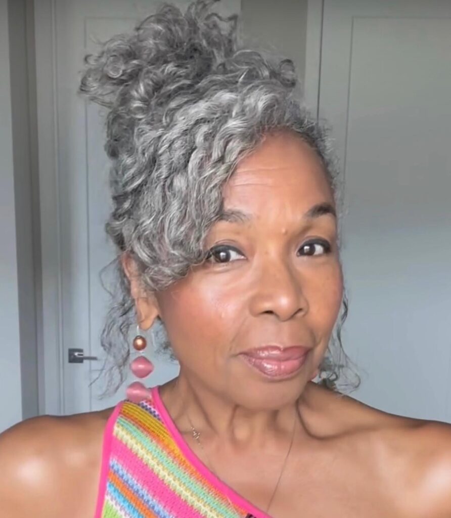 A woman with a long face shape wearing a high, curly updo in a salt-and-pepper color with curls cascading over her forehead.