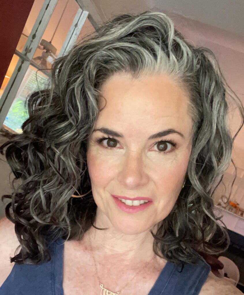 A woman with a long face shape showing off voluminous dark curly hair with a prominent silver streak along the front.
