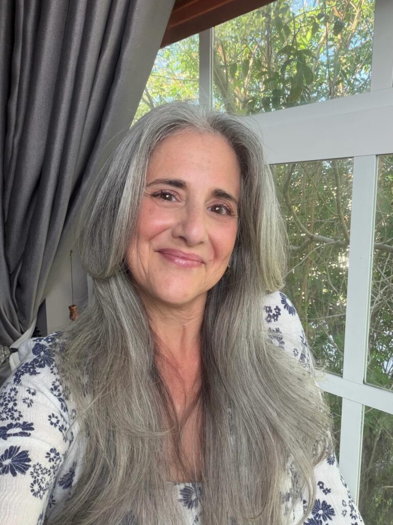 A woman with a long face shape wearing long, layered silver hair with face-framing pieces that flip outward at the cheekbones.