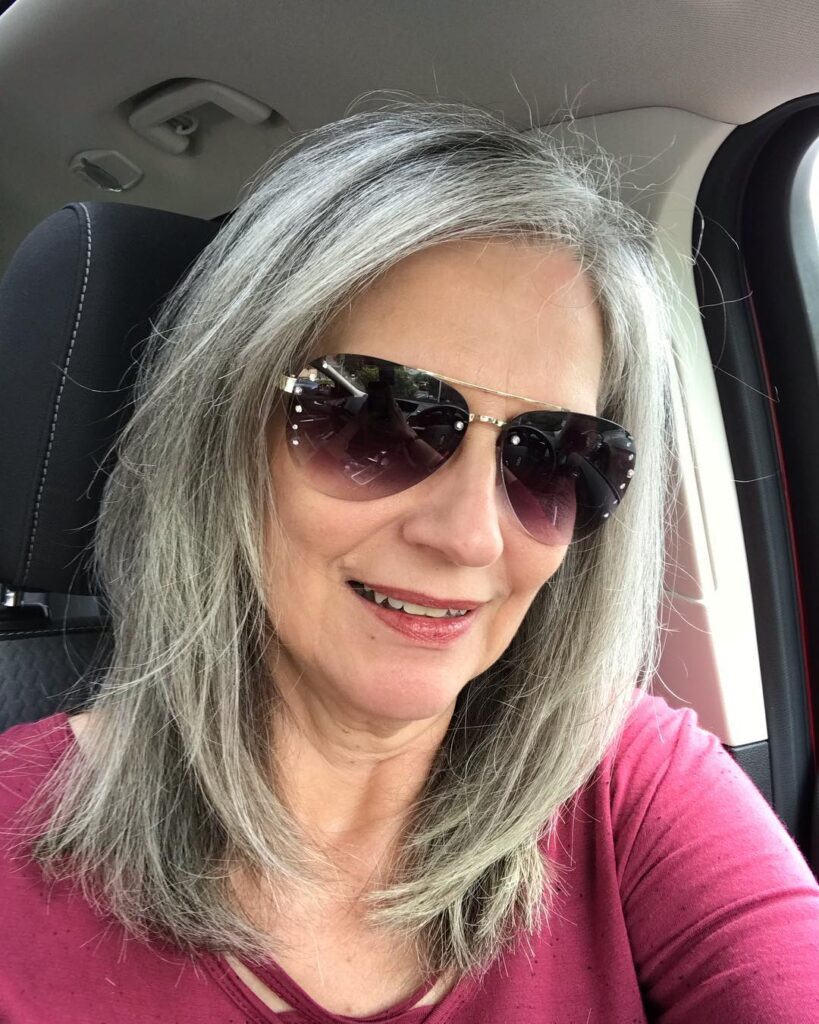 A woman wearing sunglasses and a pink top smiles while showcasing her straight, shoulder-length, silver layered hair.
