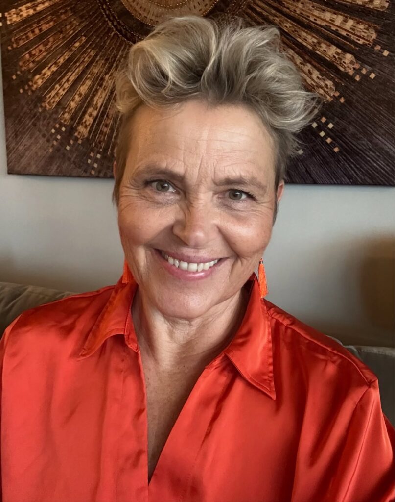 A close-up photograph of a woman with a square face shape smiling warmly at the camera, wearing a striking two-tone short pixie cut with shorter, darker sides and a voluminous, swept-up textured gray top.