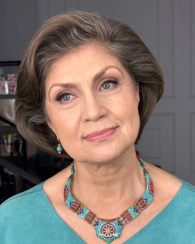 A woman over sixty with a square face shape wearing a voluminous, swept-back ashy brown and gray bob.