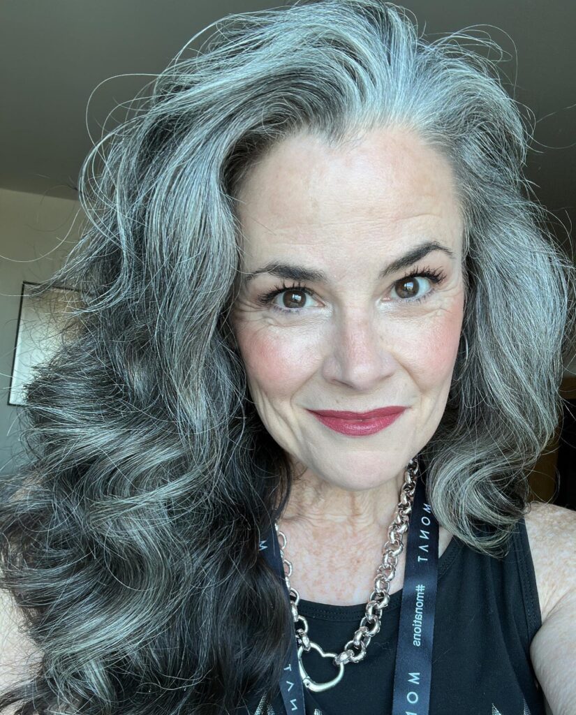 A woman with a square face and long, wavy salt-and-pepper hair styled with a soft side sweep.