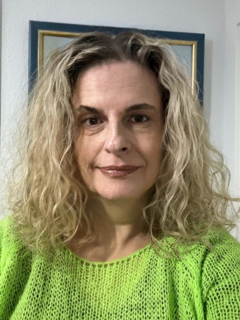 A woman over sixty with a square face shape, wearing shoulder-length sandy blonde hair styled in beachy waves with a middle part.