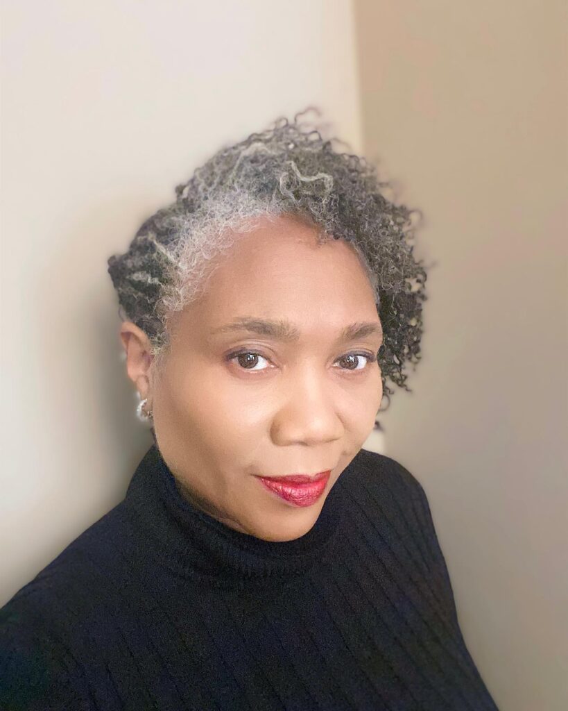 A Black woman over 60 with a tapered curly hairstyle featuring a prominent section of natural silver hair at the front.