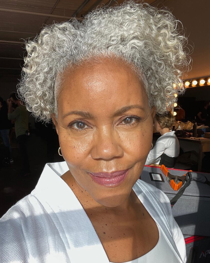 A Black woman over 60 with a voluminous halo of silver natural curls and a bright, radiant complexion.
