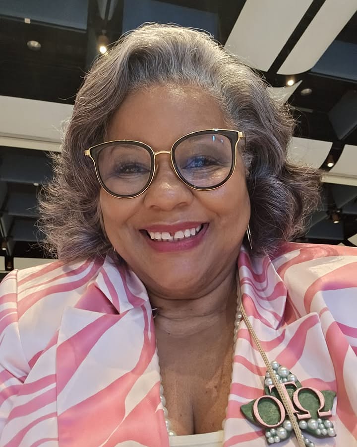 A smiling woman over 60 with a wavy silver bob hairstyle wearing black and gold glasses.