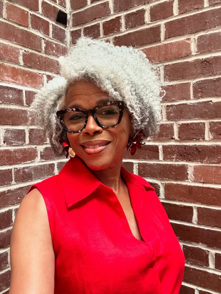 A smiling Black woman over 60 with a voluminous snow-white curly hairstyle and stylish tortoiseshell glasses.